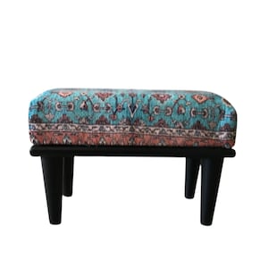 A colorful ottoman with a patterned design sits on a black wooden stand against a white background.