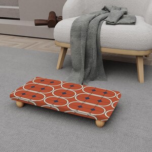 Puede incluir: Un reposapiés rectangular con un diseño moderno de mediados de siglo. La parte superior presenta un patrón repetitivo de óvalos naranjas con contornos blancos y acentos de diamantes azul marino. Se apoya en cuatro patas cortas y redondeadas de madera.