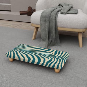 May include: A rectangular footstool featuring a teal and cream zebra stripe design. The footstool is supported by four rounded wooden legs. It rests on a grey rug. A white armchair and a grey blanket are visible in the background.