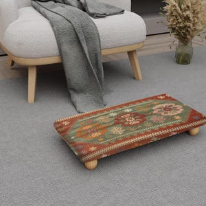 May include: A rectangular footstool with a floral pattern in shades of red, green, and orange. The footstool has four short, rounded wooden legs. The design features a central floral motif and a patterned border. The footstool is placed on a gray carpet.