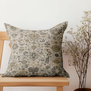 May include: A decorative throw pillow with a beige, grey, and gold patterned design. The pillow is sitting on a wooden chair.