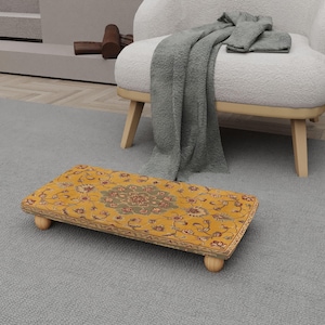 May include: A rectangular, yellow footstool with a floral pattern and wooden legs. The footstool has a detailed, ornate design with a central medallion and a border of flowers and leaves. The legs are rounded and light-colored.