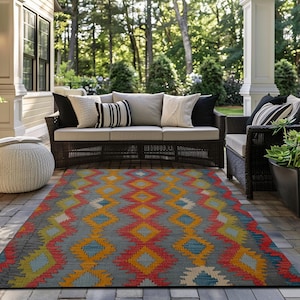 May include: A colorful outdoor rug with a geometric diamond pattern in shades of red, orange, yellow, and blue on a gray background. The rug is placed on a patio with wicker furniture and neutral-colored cushions.