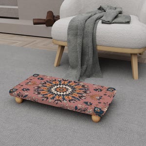 Puede incluir: Un reposapiés rectangular con un estampado floral en tonos rosa, naranja y azul. El reposapiés tiene un marco de madera y cuatro patas de madera redondeadas. El diseño recuerda a una alfombra tradicional.