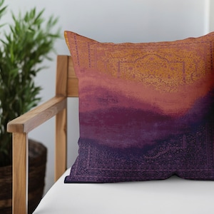 Pode incluir: Uma almofada decorativa com um design inspirado no pôr do sol, apresentando tons de laranja, vermelho, roxo e azul. A almofada tem um padrão geométrico na borda.