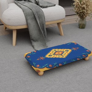 May include: A blue footstool with a decorative pattern featuring yellow, orange, and green floral elements. The footstool has a rectangular shape and is supported by four wooden legs. The design is reminiscent of a traditional rug.