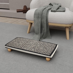 May include: A rectangular footstool with a leopard print top, black trim, and rounded wooden legs. The footstool is placed on a gray rug, with a white armchair and a gray blanket in the background.