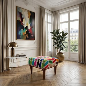 Puede incluir: Un taburete cuadrado con un patrón de hojas multicolor en tonos rojos, azules, verdes y amarillos, sostenido por cuatro patas de madera cónicas. El taburete está colocado en una habitación con una mesa auxiliar de mármol, una lámpara de latón y una pintura abstracta.