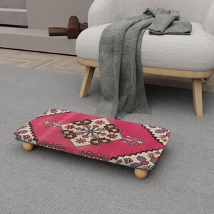 Peut inclure: Un pouf rectangulaire rose avec un motif de tapis détaillé. Le tapis présente un médaillon central avec des couleurs marron et crème, entouré de motifs floraux. Le pouf a quatre pieds en bois et est posé sur un tapis gris.