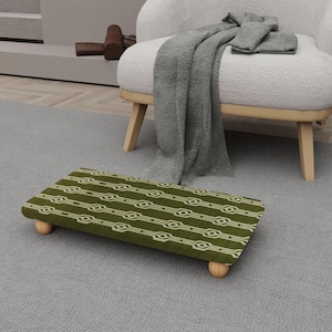 Op de afbeelding: Een rechthoekige voetenbank met een groen en wit geometrisch patroon. De voetenbank heeft vier afgeronde houten poten en staat op een grijs tapijt. Een witte fauteuil en een grijze deken staan op de achtergrond.