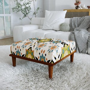 Peut inclure: Un pouf carré doté d'un revêtement en tissu crème orné d'un motif répétitif de formes florales et géométriques stylisées dans des tons bleu sarcelle, orange, vert olive et noir, reposant sur quatre pieds fuselés en bois brun moyen.