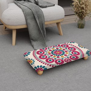 May include: A patterned footstool featuring a cream base with a circular floral design in pink, teal, yellow, and blue. The footstool, with wooden legs, sits on a grey rug. The dimensions are approximately 40 cm x 30 cm.