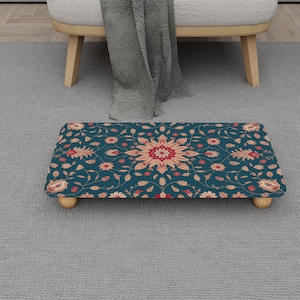 Puede incluir: Un reposapiés rectangular con un estampado floral turquesa y patas de madera. El estampado presenta un diseño floral central rojo y melocotón rodeado de flores y hojas más pequeñas. El reposapiés está colocado sobre una alfombra gris.