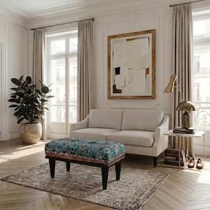 a well-furnished living room with a large window, a white sofa, a patterned ottoman, a lamp, and a plant. The room has a neutral color scheme with a touch of color from the patterned ottoman.