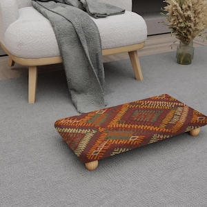 Puede incluir: Un reposapiés rectangular con una parte superior de tela estampada en tonos rojos, naranjas y verdes. El reposapiés tiene cuatro patas cortas de madera redondeadas. El reposapiés está colocado sobre una alfombra gris cerca de un sillón blanco.