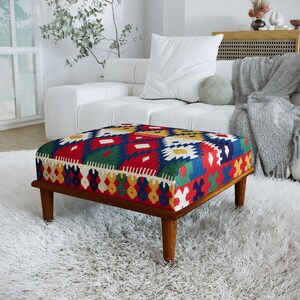 Pode incluir: Um pufe quadrado com um tecido de cores vibrantes e um padrão geométrico em vermelho, azul, verde e amarelo. A peça é sustentada por quatro pernas de madeira curtas e cónicas com um acabamento castanho natural e uma textura lisa.