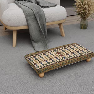 Può includere: Un poggiapiedi rettangolare con sfondo color crema e un motivo ripetuto di disegni floreali blu, rossi e verdi. Il poggiapiedi ha una struttura in legno e quattro gambe rotonde in legno. I bordi sono rifiniti con un bordo verde scuro.