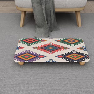 May include: A decorative footstool with a cream surface and a colourful geometric pattern in red, green, blue, and yellow. The stool has rounded corners and is supported by four small wooden legs.