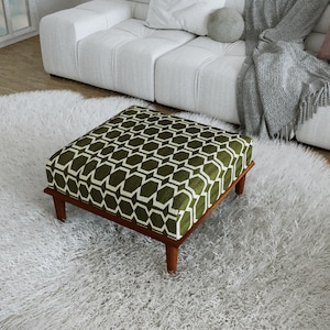 May include: A square ottoman featuring a deep olive green fabric upholstery with a repeating white geometric lattice pattern. The piece is supported by four short, tapered wooden legs in a medium brown finish, providing a stable base.