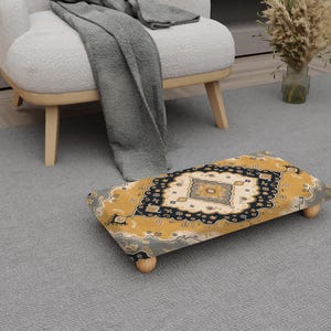 May include: A rectangular footstool with a patterned design in gold, navy, and cream. The footstool has four wooden legs and is on a grey rug. A white chair and a grey blanket are in the background.