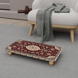 May include: A rectangular footstool with a patterned rug top in burgundy, beige, and cream colors. The footstool has four rounded wooden legs. The rug features a central medallion design and floral motifs.