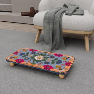 May include: A rectangular footstool with a floral design. The top features a light blue background with daisies, pink flowers, and yellow butterflies. The edges are orange, and the legs are wooden.