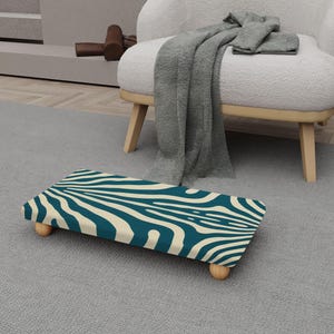 May include: A rectangular footstool with a teal and cream zebra stripe pattern. The footstool has four small, rounded wooden legs. The stool is placed on a gray rug, with a white chair and blanket in the background.