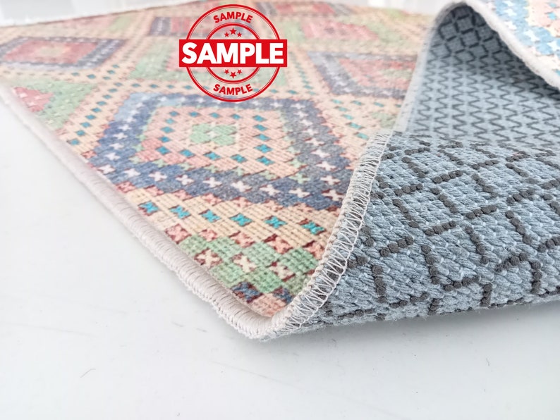 May include: A close-up of a colorful patterned rug with a gray non-slip backing. The rug has a white border and a geometric design with pink, blue, green, and yellow colors.