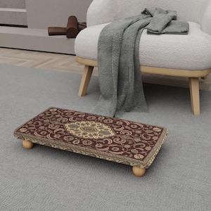 Op de afbeelding: Een rechthoekige voetenbank met een gedetailleerd, sierlijk tapijtontwerp in tinten bordeaux, beige en goud. De voetenbank heeft afgeronde houten poten en staat op een grijs tapijt. Het tapijt heeft een centraal medaillon en bloempatronen.
