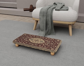 Op de afbeelding: Een rechthoekige voetenbank met een gedetailleerd, sierlijk tapijtontwerp in tinten bordeaux, beige en goud. De voetenbank heeft afgeronde houten poten en staat op een grijs tapijt. Het tapijt heeft een centraal medaillon en bloempatronen.