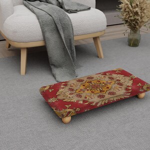 Peut inclure: Un repose-pieds rectangulaire avec un motif rouge, beige et jaune. Le repose-pieds a quatre pieds en bois et est placé sur un tapis gris. Le motif présente des éléments floraux et géométriques.