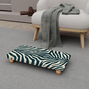 May include: A rectangular footstool with a zebra print design in teal and cream. The footstool has four rounded wooden legs. The footstool is placed on a gray rug.