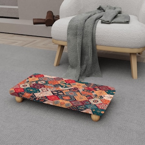 May include: A rectangular footstool with a colorful patchwork design. The pattern includes various geometric shapes in shades of red, orange, green, and blue. The footstool is supported by four small, rounded wooden legs.