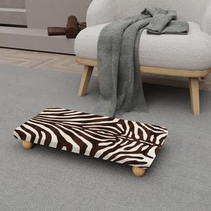 May include: A rectangular footstool with a zebra print pattern in brown and white. The footstool has four short, rounded wooden legs. The footstool is placed on a gray carpet, near a white armchair.