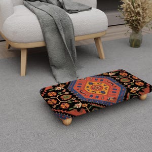 May include: A patterned footstool featuring a detailed design in red, blue, orange, and black. The footstool has short, light-coloured wooden legs and is placed on a grey carpet. The design is reminiscent of a traditional rug.