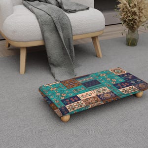 May include: A rectangular footstool with a patchwork design in shades of teal, navy, and brown. The stool has four small, rounded wooden legs. The patchwork pattern features geometric and floral motifs. The footstool is placed on a gray carpet.