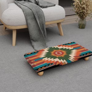 May include: A rectangular footstool with a colorful geometric pattern in shades of orange, teal, green, and cream. The footstool has a black border and is supported by four small, rounded wooden legs. The design is reminiscent of Native American art.