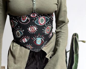May include: A black corset with a pattern of colorful eyes. The corset is worn over a green long-sleeved shirt.