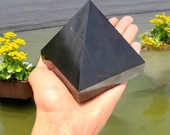 May include: A polished black pyramid made of stone. The pyramid is held in an open hand, with a water background and yellow flowers in the background. The pyramid has a smooth, reflective surface.