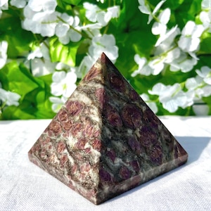 May include: A pyramid-shaped decorative object made of a stone with a mix of burgundy and gray tones. The pyramid has a square base and tapers to a point at the top. The stone has a textured surface with a pattern of burgundy spots and gray veins.