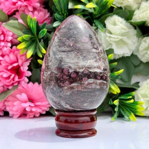 Peut inclure: Une sculpture en pierre polie en forme d'œuf présentant une base marbrée gris foncé et blanc avec des cristaux de grenat rouge profond incrustés. La pierre est montée sur un socle circulaire en bois brun foncé, avec une finition lisse et brillante.