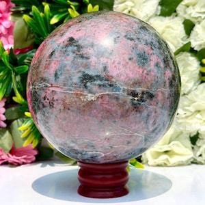 May include: A polished spherical stone featuring a mottled pattern of dusty pink, deep grey, and black mineral inclusions. The sphere rests securely on a small, circular, dark reddish-brown wooden stand, showcasing a smooth, reflective surface.