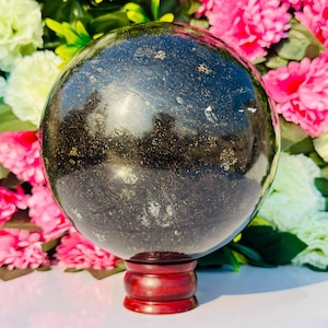 Op de afbeelding: Een grote, gepolijste zwarte bol met gouden vlekken, rustend op een donkerrode houten standaard. De bol reflecteert licht, waardoor een glinsterend effect ontstaat. Roze en witte bloemen op de achtergrond.