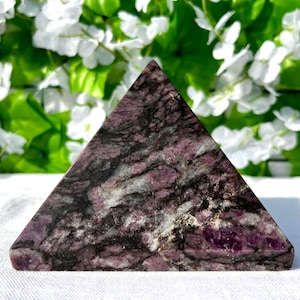 Könnte beinhalten: Ein pyramidenförmiger Stein mit einer Mischung aus dunkelvioletten, schwarzen und weißen Adern. Die Pyramide befindet sich vor weißen Blumen und grünen Blättern. Die Oberfläche des Steins ist poliert.