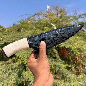 Puede incluir: Un cuchillo de obsidiana negra con una hoja dentada y un mango envuelto en hilo beige. El cuchillo se sostiene sobre un fondo de follaje verde y un cielo azul claro.