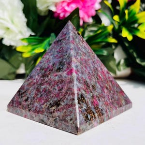 Könnte beinhalten: Ein pyramidenförmiges Dekorationsobjekt aus Stein mit einer Mischung aus grauen, schwarzen und rosa Farbtönen. Die Pyramide hat eine glatte, polierte Oberfläche und ein symmetrisches Design. Der Hintergrund zeigt bunte Blumen.