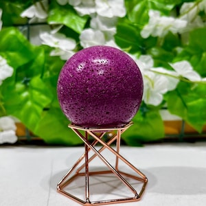 Könnte beinhalten: Eine tiefviolette Kugel mit strukturierter Oberfläche, präsentiert auf einem geometrischen roségoldenen Metallständer. Die Kugel ist der Mittelpunkt, vor einem Hintergrund aus grünen Blättern und weißen Blüten. Der Ständer hat ein modernes, offenes Design.