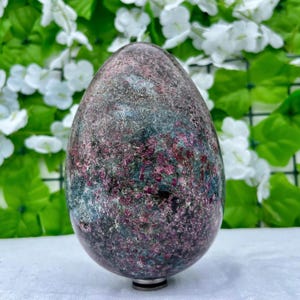 Pode incluir: Uma pedra grande em forma de ovo com uma aparência manchada. A pedra apresenta uma mistura de cores cinza escuro, vermelho profundo e verde-azulado. A superfície polida reflete a luz, destacando as texturas e tons variados da pedra natural. A base tem um anel de metal prateado.