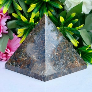May include: A polished pyramid sculpture crafted from dark brown and black stone with visible crystalline textures and natural mineral inclusions. The object features smooth, flat triangular faces that meet at a sharp apex, showcasing a glossy finish.