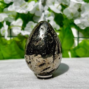 May include: A pyrite egg-shaped sculpture with a polished finish. The surface has a metallic sheen with areas of dark and light coloration. The egg sits on a small, round base with a black and white ring. The background features white flowers and green foliage.